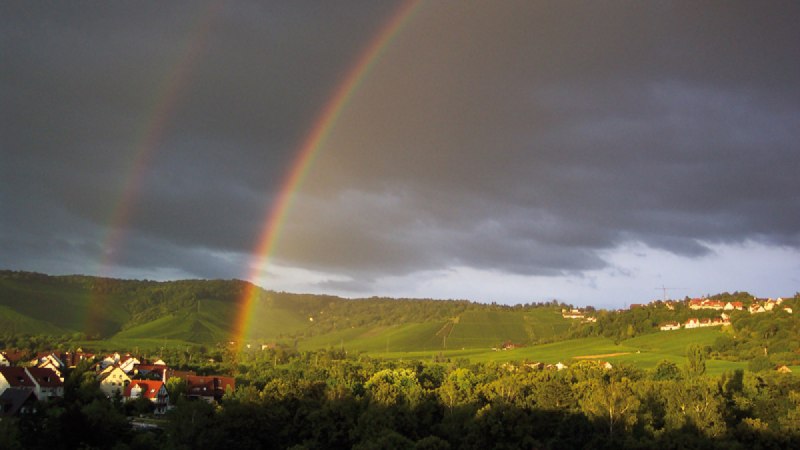 Doppelter Regenbogen über grüner Landschaft mit Häusern, dunklen Wolken und Sonnenlicht auf den Hügeln., © Stuttgart-Marketing GmbH