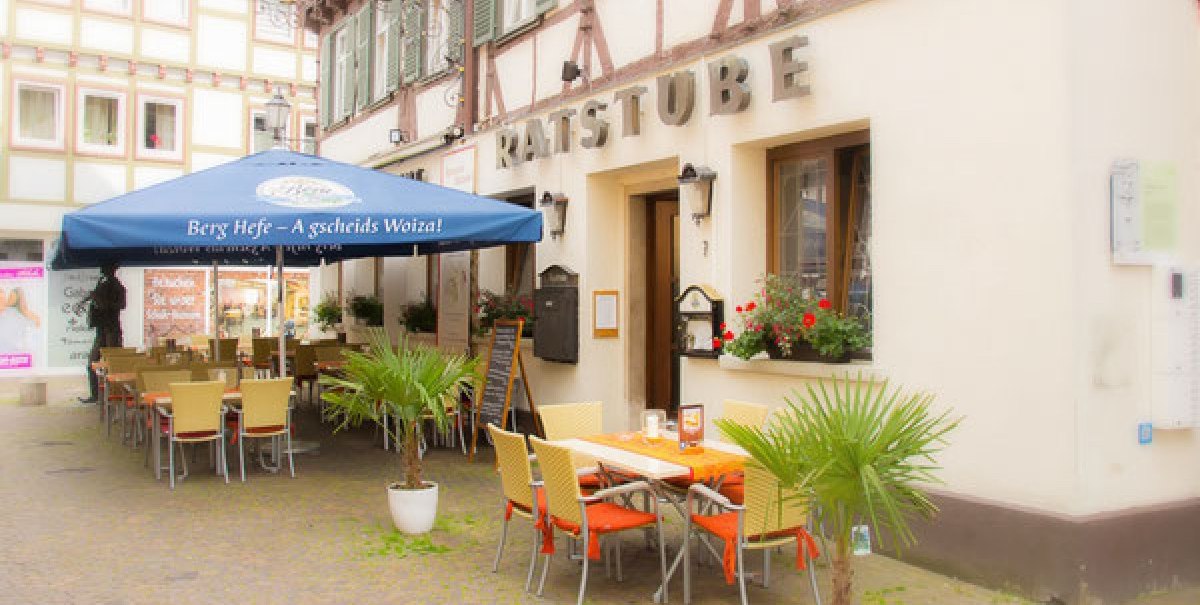 Ein Restaurant namens Ratsstube mit Außenbestuhlung unter einem blauen Sonnenschirm. Fachwerkgebäude im Hintergrund, Pflanzen schmücken den Eingangsbereich.