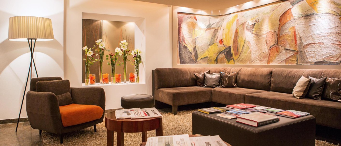 Moderne Lobby mit braunem Sofa, abstraktem Kunstwerk, Stehlampe und Zeitschriften auf einem Tisch. Blumen in Vasen dekorieren den Raum., © Ringhotel Adler Asperg