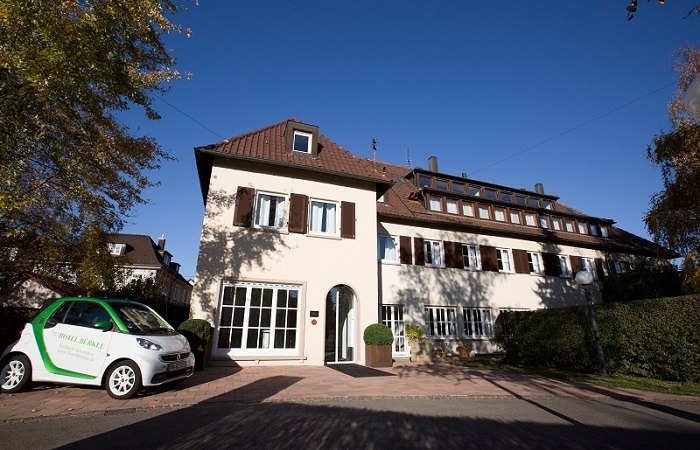 Ein zweistöckiges Hotel mit braunen Fensterläden, umgeben von Bäumen. Ein kleines Auto steht davor. Der Himmel ist klar und blau., © Hotel Bürkle Ein zweistöckiges Hotel mit braunen Fensterläden, umgeben von Bäumen. Ein kleines Auto steht davor. Der Himmel ist klar und blau., © Hotel Bürkle