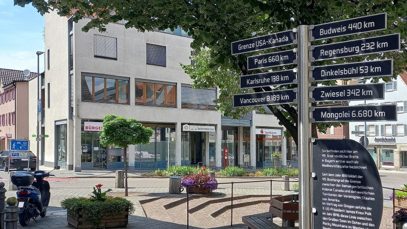 Wegweiser auf einem Platz mit Entfernungen zu St&auml;dten und Orten auf dem 49. Breitengrad, wie Paris und Vancouver. Im Hintergrund ein modernes, wei&szlig;es Geb&auml;ude, &copy; Petra Natzkowski