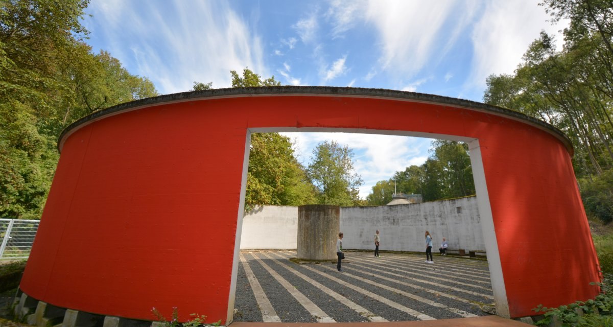 Rote Betonwand mit großem rechteckigem Durchgang, dahinter Menschen auf gestreiftem Boden. Umgeben von Bäumen und blauem Himmel., © Fellbach Tourismus Rote Betonwand mit großem rechteckigem Durchgang, dahinter Menschen auf gestreiftem Boden. Umgeben von Bäumen und blauem Himmel., © Fellbach Tourismus