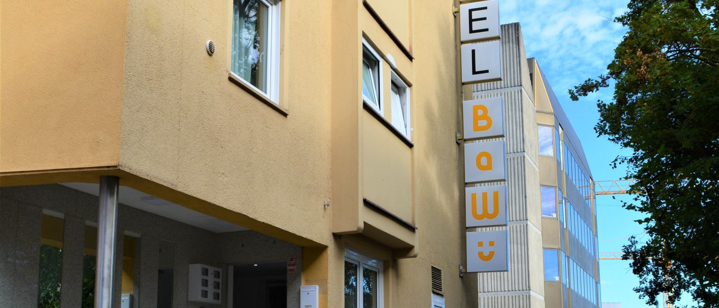 Gelbes Hotelgebäude mit Schild 'Hotel BaWü', Treppe zum Eingang, umgeben von Bäumen und modernen Gebäuden im Hintergrund., © Hotel BaWü