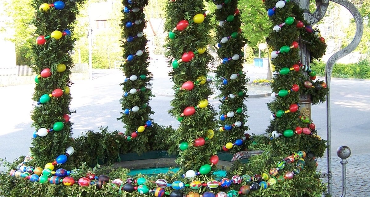 Ein traditionell geschmückter Osterbrunnen mit bunten Eiern und grünen Girlanden, umgeben von Bäumen und gepflastertem Boden., © Natur.Nah. Schönbuch & Heckengäu Ein traditionell geschmückter Osterbrunnen mit bunten Eiern und grünen Girlanden, umgeben von Bäumen und gepflastertem Boden., © Natur.Nah. Schönbuch & Heckengäu