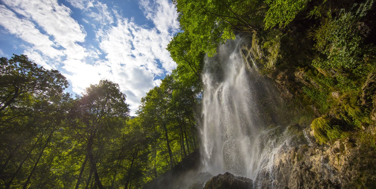 Der Uracher Wasserfall stürzt über Felsen, umgeben von dichtem Wald. Der Himmel ist blau mit weißen Wolken., © Bad Urach Tourismus