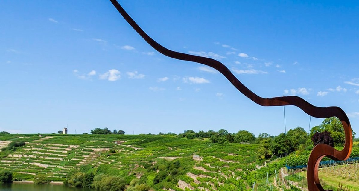 Weinberge in Kirchheim unter blauem Himmel, mit einer geschwungenen Skulptur im Vordergrund. Ein See und grüne Landschaft im Hintergrund., © Land der 1000 Hügel - Kraichgau-Stromberg Weinberge in Kirchheim unter blauem Himmel, mit einer geschwungenen Skulptur im Vordergrund. Ein See und grüne Landschaft im Hintergrund., © Land der 1000 Hügel - Kraichgau-Stromberg