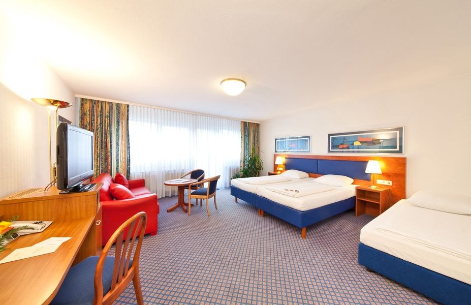 Geräumiges Hotelzimmer mit zwei Betten, rotem Sofa, Schreibtisch, Fernseher und großem Fenster mit Vorhängen., © Novum Boulevard Stuttgart City