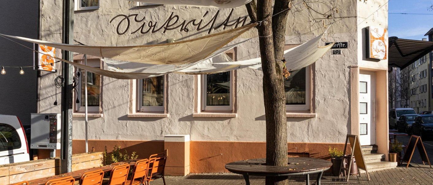 Ein Caf&eacute; mit Sonnensegeln und orangenen St&uuml;hlen im Au&szlig;enbereich. Die Fassade ist beige mit Schriftzug. Ein Baum steht vor dem Eingang., &copy; Stuttgart-Marketing GmbH, Sarah Schmid