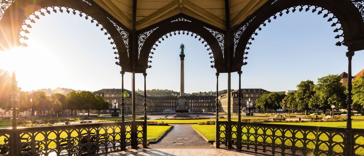 Blick durch einen Pavillon auf den Schlossplatz Stuttgart mit der Jubiläumssäule, umgeben von historischen Gebäuden und grünen Rasenflächen im Sonnenlicht., © Stuttgart-Marketing GmbH Werner Dieterich