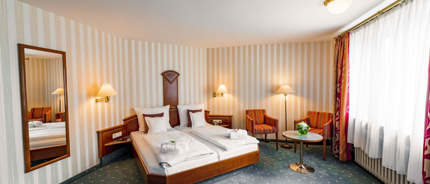 Gemütliches Hotelzimmer mit Doppelbett, zwei Sesseln, rundem Tisch und großen Fenstern mit Vorhängen. Elegante Beleuchtung und gestreifte Tapete., © TOMAS
