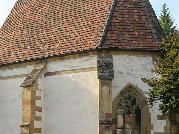 Historische Kirche mit rotem Ziegeldach, gotischem Fenster und Tauben auf dem Dach. Ein Baum und ein Schild sind ebenfalls sichtbar., © Alex Becher