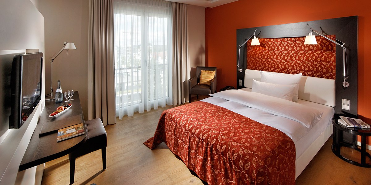 Elegantes Hotelzimmer mit roter Akzentwand, Doppelbett, Schreibtisch, Fernseher und großem Fenster mit Vorhängen., © Parkhotel Stuttgart Messe-Airport