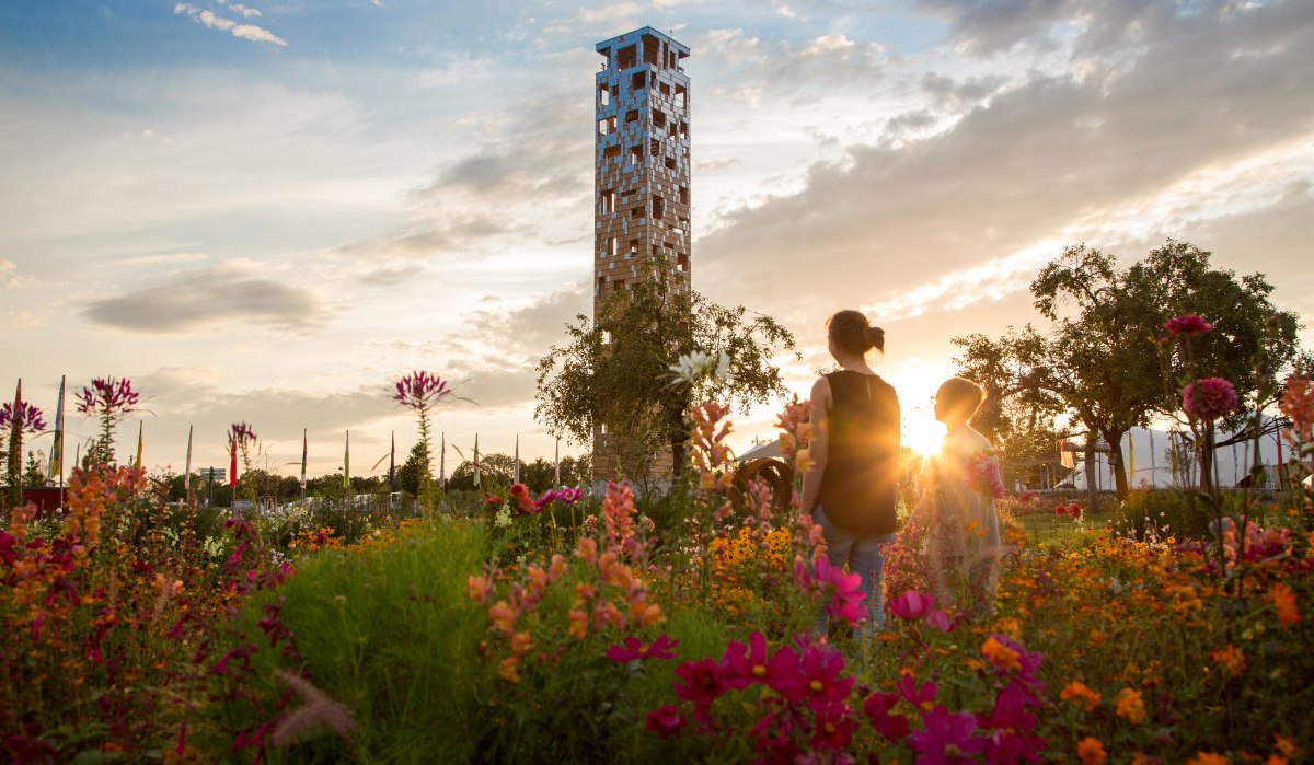 Ein Turm im Himmelsgarten, umgeben von bunten Blumen. Zwei Personen stehen im Vordergrund, während die Sonne im Hintergrund untergeht., © Foto Thomas Zehnder
