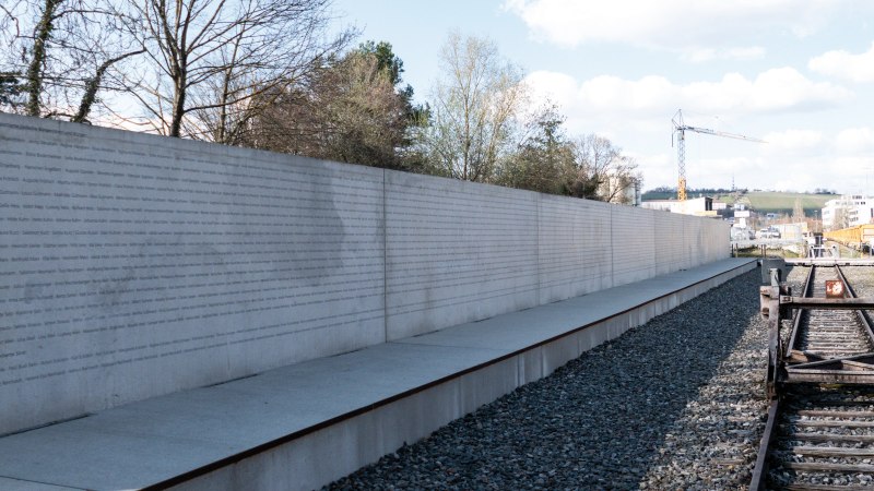 Eine lange Betonwand mit eingravierten Namen steht neben stillgelegten Bahngleisen unter einem bewölkten Himmel., © Foto-Kunst Andreas Keller