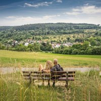 Ein Paar sitzt auf einer Bank und blickt auf eine grüne Landschaft mit einem Dorf im Hintergrund., © Natur.Nah. Schönbuch & Heckengäu Ein Paar sitzt auf einer Bank und blickt auf eine grüne Landschaft mit einem Dorf im Hintergrund., © Natur.Nah. Schönbuch & Heckengäu