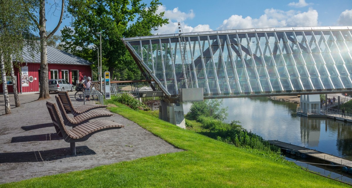 Moderne Glasbrücke über Fluss in Remseck am Neckar, umgeben von grüner Landschaft und einem roten Gebäude. Sitzbänke und Radfahrer im Vordergrund., © Remstal Tourismus e.V.