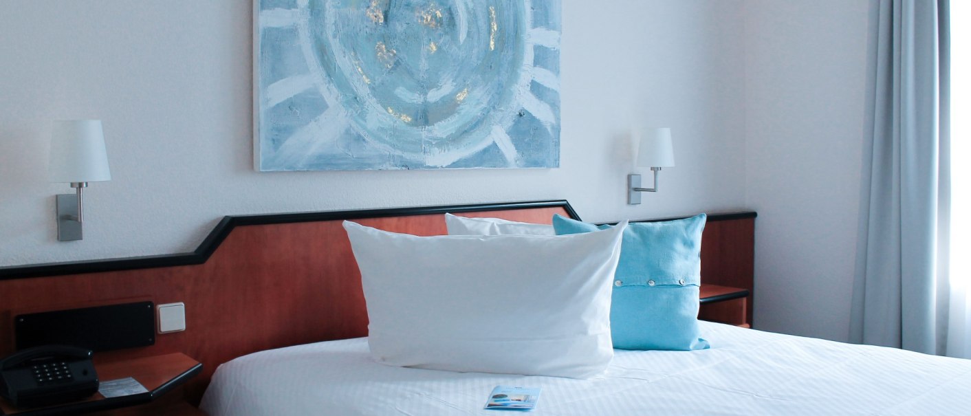 Hotelzimmer mit Doppelbett, blauen Kissen, abstraktem Gemälde und Nachttischen mit Lampen., © Hotel Princess