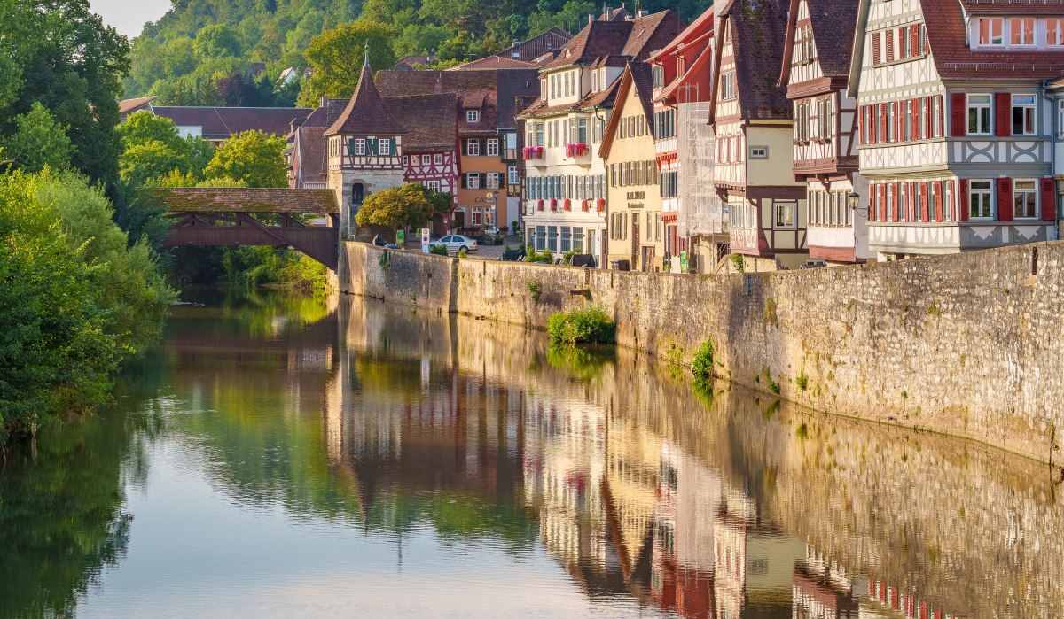 Fachwerkhäuser in Schwäbisch Hall spiegeln sich im Fluss. Eine Brücke und grüne Hügel im Hintergrund vervollständigen die malerische Szene., © Michael Kühneisen Fachwerkhäuser in Schwäbisch Hall spiegeln sich im Fluss. Eine Brücke und grüne Hügel im Hintergrund vervollständigen die malerische Szene., © Michael Kühneisen