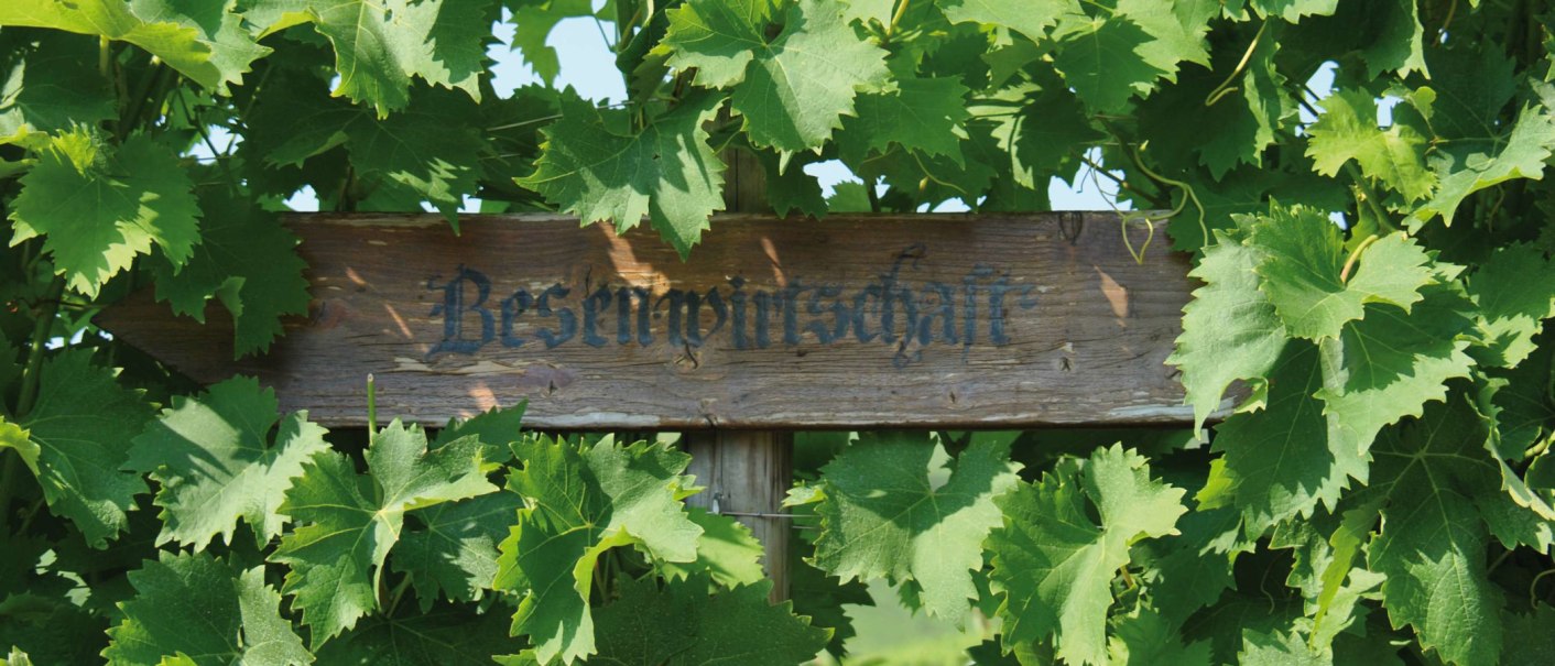 Ein Holzschild mit der Aufschrift 'Besenwirtschaft' ist von grünen Weinblättern umgeben., © SMG