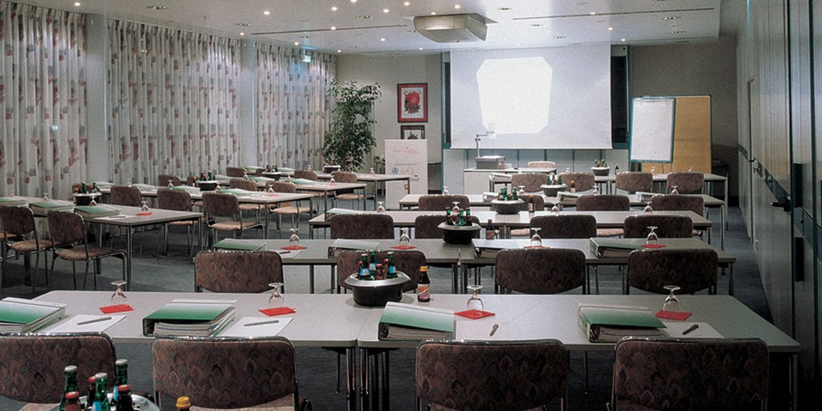 Ein leerer Konferenzraum mit Tischen, Stühlen, einem Projektor und einem Flipchart. Auf den Tischen liegen Ordner und Wasserflaschen., © © H-Hotels.com