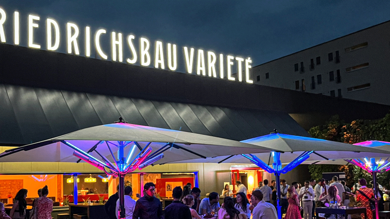 Abendliche Szene vor dem Friedrichsbau Varieté mit beleuchteten Sonnenschirmen und Menschen, die sich unterhalten und entspannen., © Friedrichsbau Varieté Abendliche Szene vor dem Friedrichsbau Varieté mit beleuchteten Sonnenschirmen und Menschen, die sich unterhalten und entspannen., © Friedrichsbau Varieté