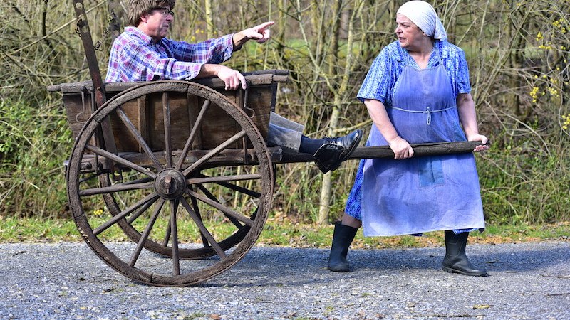 Eine Frau in Sch&uuml;rze zieht einen Mann in einem Holzwagen. Beide tragen l&auml;ndliche Kleidung und befinden sich auf einem Weg im Freien., &copy; EMT Event-Media-Tec GmbH