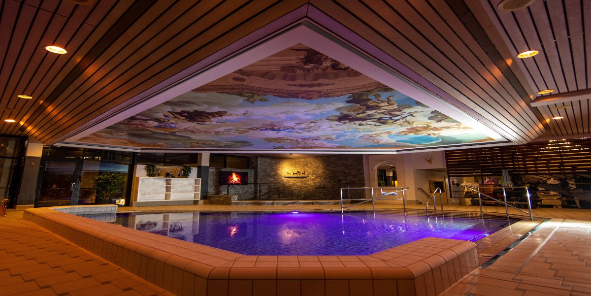 Ein beleuchteter Innenpool in den AlbThermen mit einer kunstvoll bemalten Decke und moderner Einrichtung., © AlbThermen Bad Urach