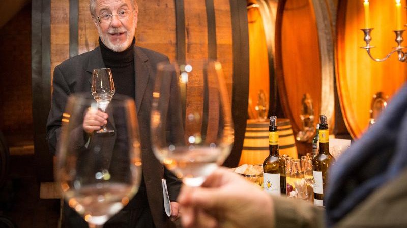 Ein Mann in einem Weinkeller hält ein Weinglas. Im Hintergrund sind große Holzfässer und Kerzenleuchter zu sehen., © © T&E, Benjamin Stollenberg Ein Mann in einem Weinkeller hält ein Weinglas. Im Hintergrund sind große Holzfässer und Kerzenleuchter zu sehen., © © T&E, Benjamin Stollenberg