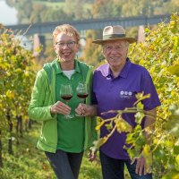 Zwei Personen genie&szlig;en Rotwein in einem sonnigen Weinberg. Sie l&auml;cheln und halten Weingl&auml;ser. Im Hintergrund ist eine Br&uuml;cke sichtbar., &copy; &copy; Hilde Hampp