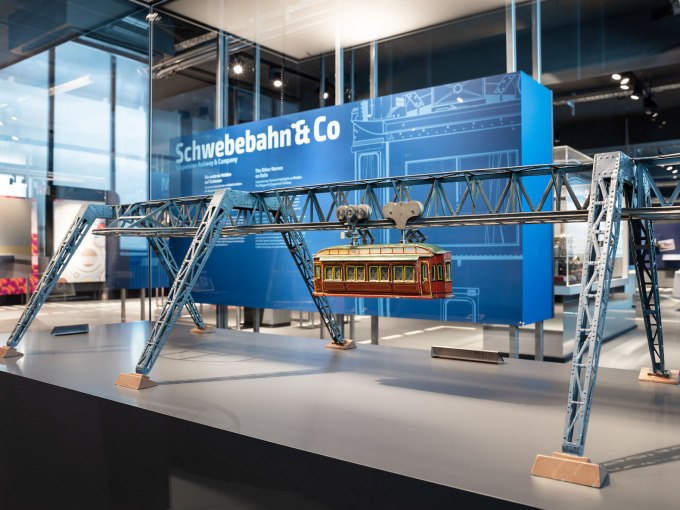 Ein Modell einer Schwebebahn im M&auml;rklineum, ausgestellt auf einem Tisch. Im Hintergrund ist ein blauer Infostand zu sehen., &copy; M&auml;rklin Spielwaren Vertriebs-GmbH