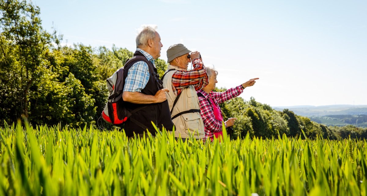 Drei ältere Personen wandern durch ein grünes Feld, zeigen in die Ferne und genießen die Natur. Sie tragen Freizeitkleidung und Rucksäcke., © Distretto di Göppingen Drei ältere Personen wandern durch ein grünes Feld, zeigen in die Ferne und genießen die Natur. Sie tragen Freizeitkleidung und Rucksäcke., © Distretto di Göppingen