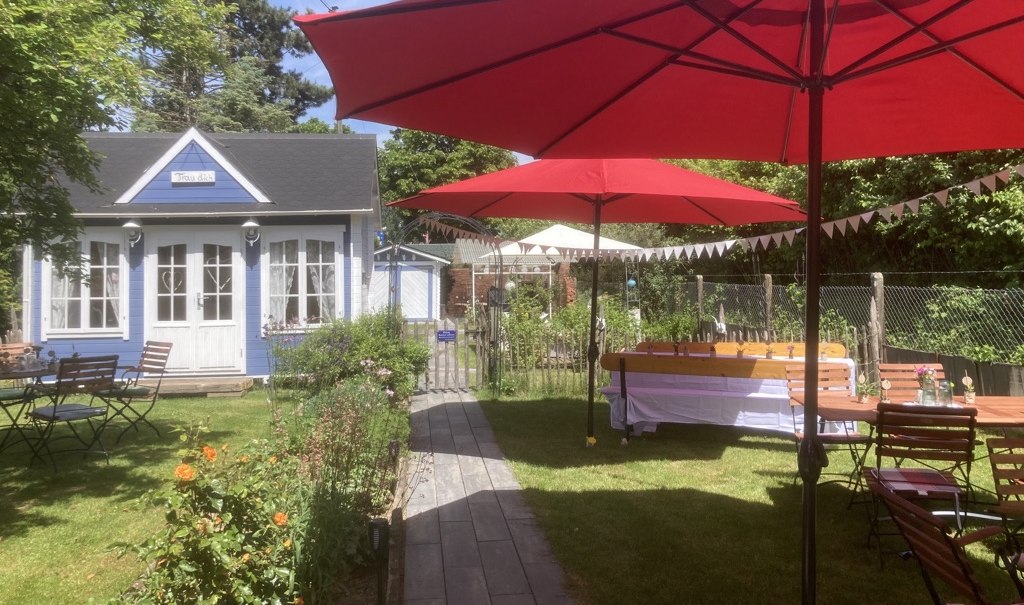 Garten Café mit roten Sonnenschirmen, Holztischen und -stühlen, umgeben von Blumen und Bäumen. Ein kleines blaues Gebäude im Hintergrund., © Café Plätschwiesle