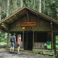 Zwei Personen betrachten die Rolf Hamann Hütte im Wald. Die Hütte ist aus Holz und von Bäumen umgeben., © LOCHER Fotodesign&Manufaktur