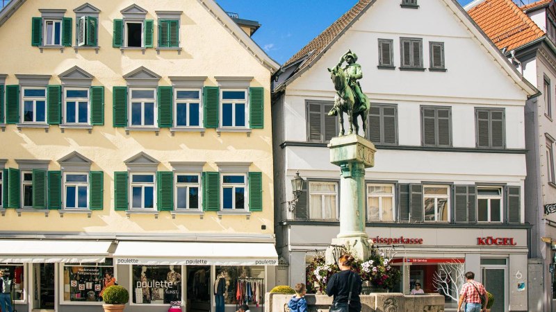 Der Postmichelbrunnen auf dem Marktplatz in Esslingen, flankiert von historischen Gebäuden mit Geschäften im Erdgeschoss. Menschen spazieren vorbei., © Stuttgart-Marketing GmbH, Sarah Schmid
