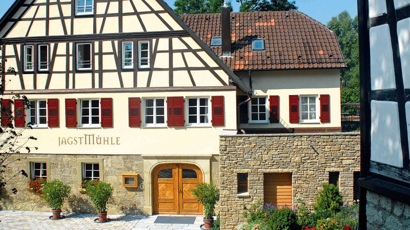 Fachwerkhaus mit rotem Dach und Fensterl&auml;den, beschriftet mit 'Jagstm&uuml;hle'. Vor dem Haus stehen Pflanzen in T&ouml;pfen., &copy; Landgasthof + Hotel Jagstm&uuml;hle