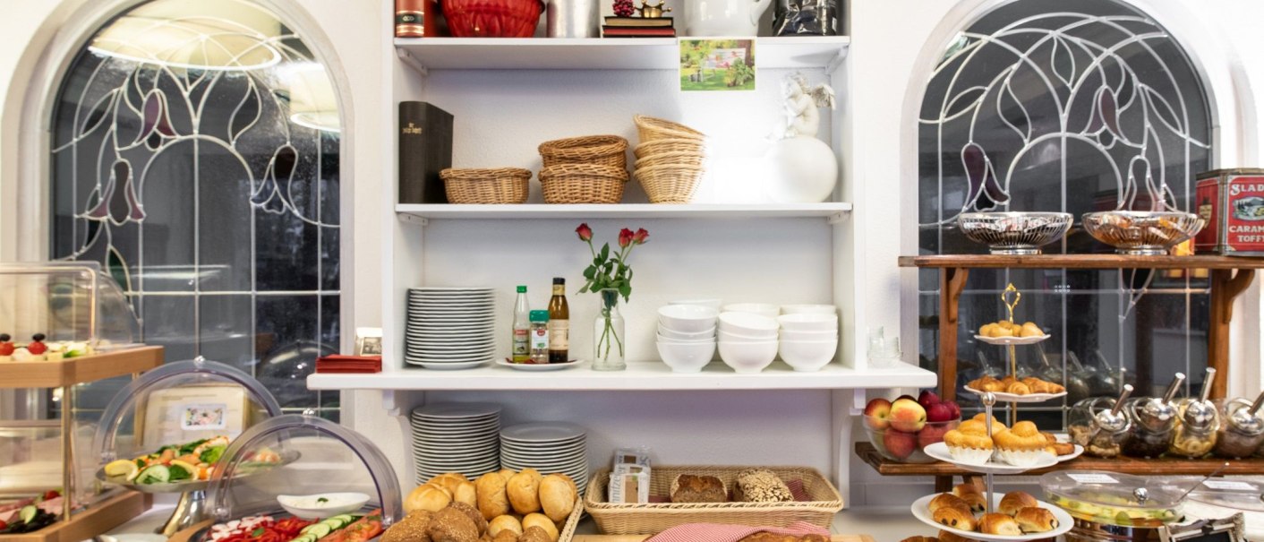Ein reichhaltiges Frühstücksbuffet mit Brot, Gebäck, Obst und Geschirr. Im Hintergrund sind dekorative Fenster zu sehen., © Hotel Weinstadt
