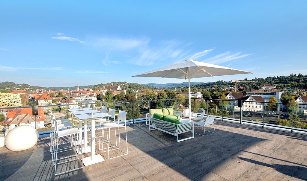 Moderne Dachterrasse mit weißen Möbeln, Sonnenschirm und Blick auf eine Stadtlandschaft mit Hügeln im Hintergrund., © Hotel am Remspark Moderne Dachterrasse mit weißen Möbeln, Sonnenschirm und Blick auf eine Stadtlandschaft mit Hügeln im Hintergrund., © Hotel am Remspark