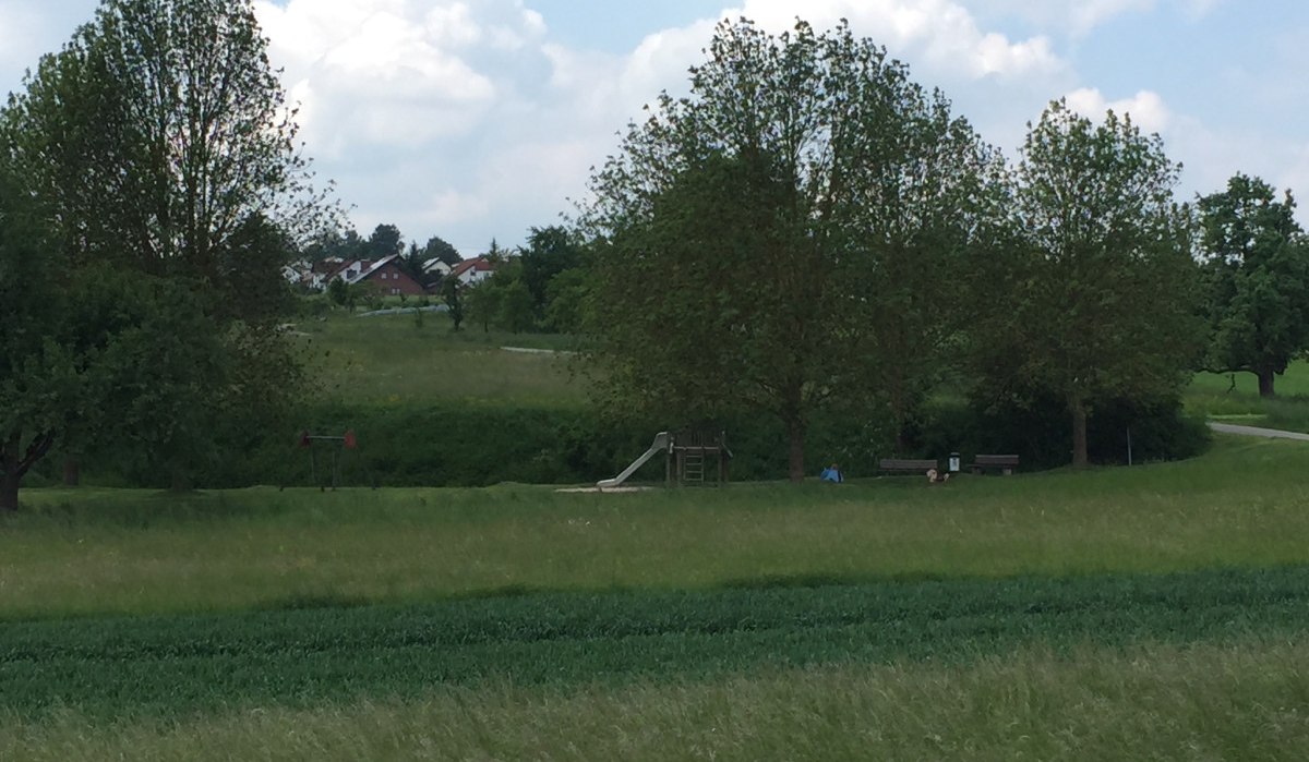 Ein Spielplatz mit Rutsche und Bänken in einer grünen Wiesenlandschaft, umgeben von Bäumen. Im Hintergrund sind Häuser zu sehen., © www.pro-cycl.de Ein Spielplatz mit Rutsche und Bänken in einer grünen Wiesenlandschaft, umgeben von Bäumen. Im Hintergrund sind Häuser zu sehen., © www.pro-cycl.de