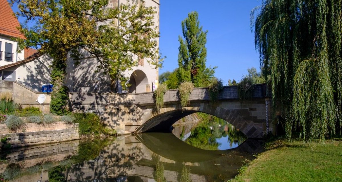 Der Beinsteiner Torturm steht neben einer Brücke, die sich im ruhigen Wasser spiegelt. Der Himmel ist klar und blau, umgeben von Bäumen., © WTM GmbH Waiblingen Der Beinsteiner Torturm steht neben einer Brücke, die sich im ruhigen Wasser spiegelt. Der Himmel ist klar und blau, umgeben von Bäumen., © WTM GmbH Waiblingen