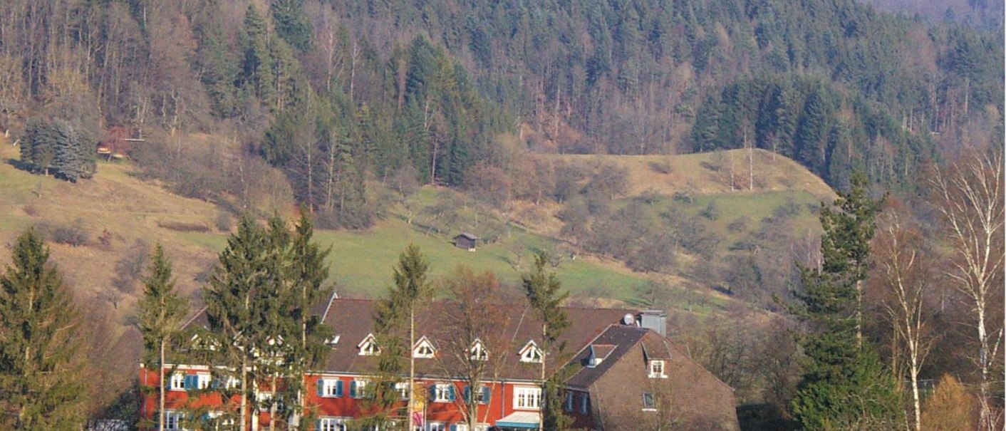 Ein rotes Gebäude mit weißen Fenstern steht vor einem bewaldeten Hügel. Im Vordergrund sind Bäume und Sträucher zu sehen., © DJH Baden-Württemberg e.V.