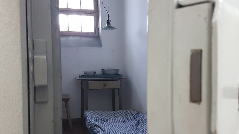 Blick in eine historische Gef&auml;ngniszelle mit einfachem Bett, kleinem Tisch und Sch&uuml;sseln. Ein Fenster mit Gitterst&auml;ben l&auml;sst Licht herein., &copy; Strafvollzugsmuseum Ludwigsburg