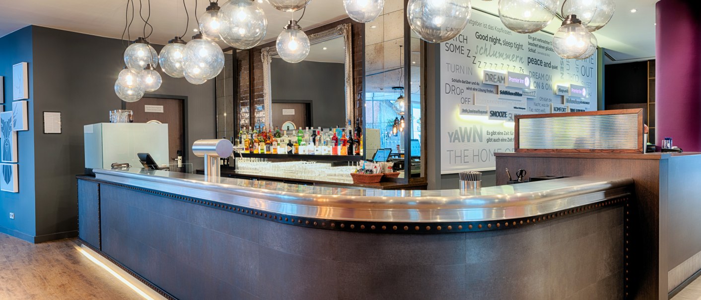 Moderne Bar mit geschwungener Theke, hängenden Glaskugellampen und einem Regal voller Flaschen im Hintergrund., © Premier Inn Germany Moderne Bar mit geschwungener Theke, hängenden Glaskugellampen und einem Regal voller Flaschen im Hintergrund., © Premier Inn Germany