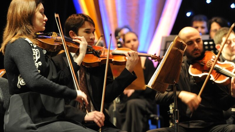 Ein Orchester spielt auf einer Bühne. Mehrere Musiker sind mit Geigen zu sehen, konzentriert auf ihre Instrumente. Hintergrund mit bunten Lichtern., © MCS Ein Orchester spielt auf einer Bühne. Mehrere Musiker sind mit Geigen zu sehen, konzentriert auf ihre Instrumente. Hintergrund mit bunten Lichtern., © MCS