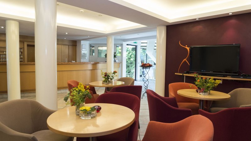 Moderne Hotel-Lobby mit gem&uuml;tlichen Sesseln, Tischen mit Blumen, einem gro&szlig;en Fernseher und einer Rezeption im Hintergrund., &copy; Hotel Gloria GmbH Restaurant M&ouml;hringer Hexle