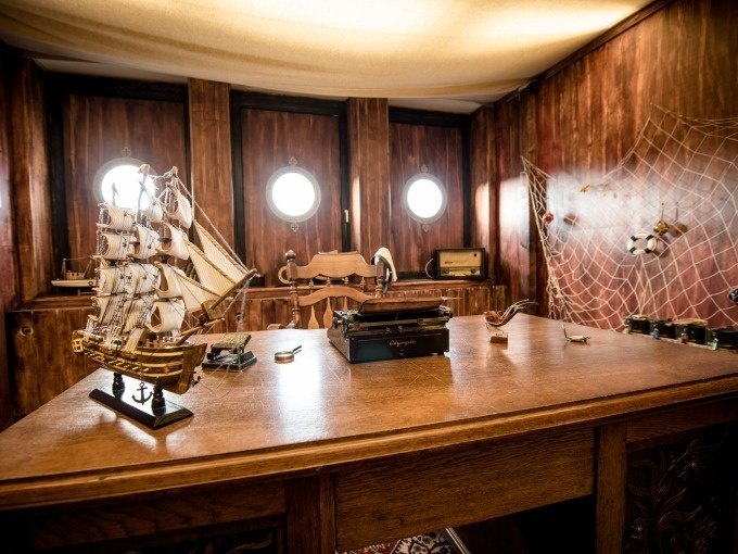Ein hölzerner Schreibtisch mit Modellschiff und Schreibmaschine in einem maritimen Raum. An der Wand hängt ein Fischernetz., © ExitGames Stuttgart Ein hölzerner Schreibtisch mit Modellschiff und Schreibmaschine in einem maritimen Raum. An der Wand hängt ein Fischernetz., © ExitGames Stuttgart