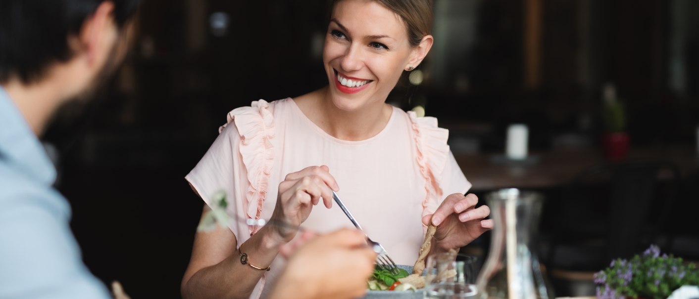 Eine Frau in einem Restaurant lächelt beim Essen, während sie mit einem Mann spricht. Auf dem Tisch stehen Gläser und eine Karaffe., © Envato Elements Eine Frau in einem Restaurant lächelt beim Essen, während sie mit einem Mann spricht. Auf dem Tisch stehen Gläser und eine Karaffe., © Envato Elements