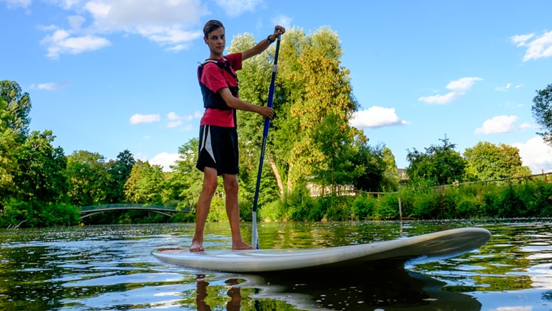 Eine Person steht auf einem Stand Up Paddle Board auf einem Fluss, umgeben von grüner Natur und blauem Himmel., © WTM GmbH Waiblingen Eine Person steht auf einem Stand Up Paddle Board auf einem Fluss, umgeben von grüner Natur und blauem Himmel., © WTM GmbH Waiblingen