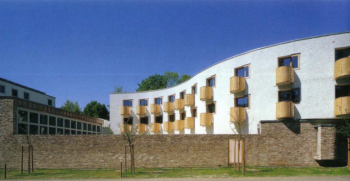 Modernes Gebäude mit wellenförmiger Fassade und runden Balkonen, umgeben von einer Steinmauer, vor blauem Himmel., © TOMAS