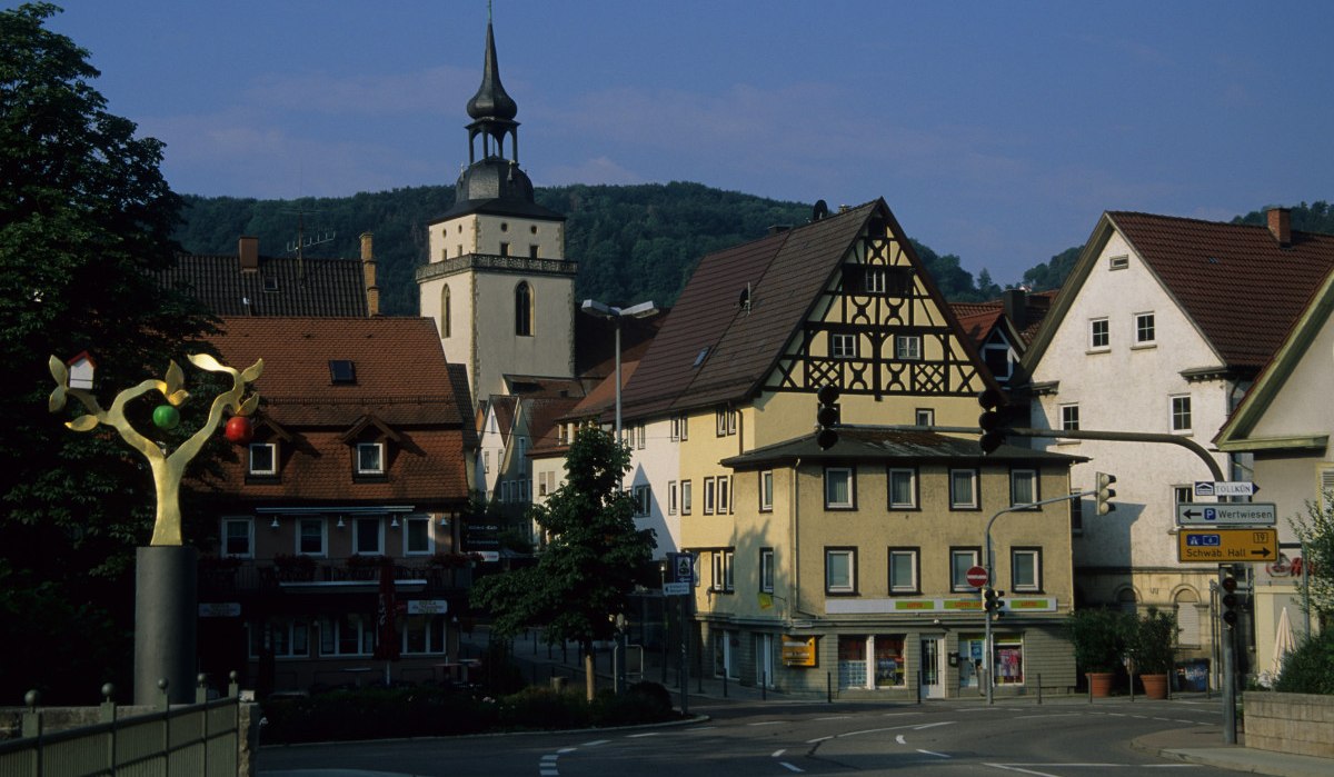 Stadtansicht von Künzelsau mit Fachwerkhäusern, einem Kirchturm und einer modernen Skulptur im Vordergrund. Straßenschilder und Ampeln sind sichtbar., © Touristikgemeinschaft Hohenlohe, Künzelsau / Eva Maria Kraiss Stadtansicht von Künzelsau mit Fachwerkhäusern, einem Kirchturm und einer modernen Skulptur im Vordergrund. Straßenschilder und Ampeln sind sichtbar., © Touristikgemeinschaft Hohenlohe, Künzelsau / Eva Maria Kraiss