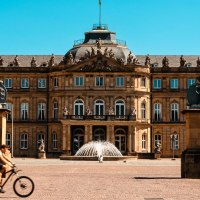 Das Neue Schloss Stuttgart bei sonnigem Wetter. Menschen gehen und ein Fahrradfahrer f&auml;hrt im Vordergrund. Zwei Statuen flankieren den Eingang., &copy; Stuttgart-Marketing GmbH, Sarah Schmid